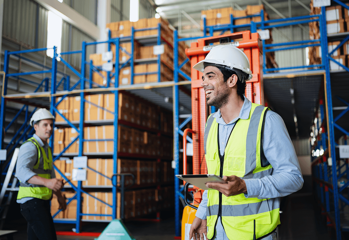 Employee with clipboard in warehouse aisle showing inventory slotting and stacked boxes.
